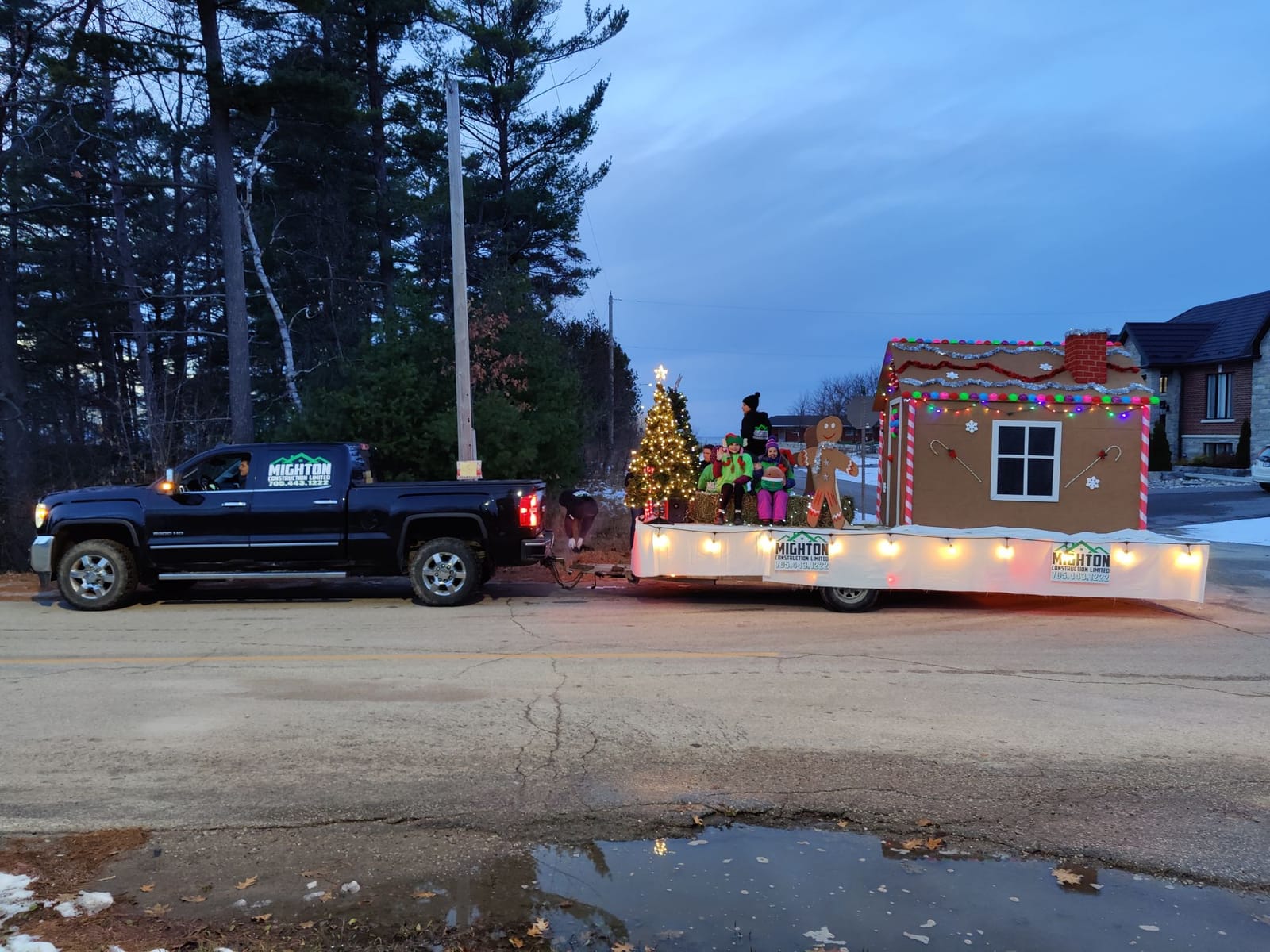 Mighton Construction gingerbread-house float with Christmas lights being towed by a pickup truck in the 2023 Wasaga Beach Santa Claus Parade